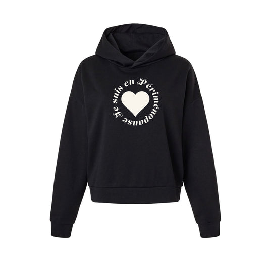 A black cropped pullover hoodie with a white graphic on the chest featuring a heart and circular text JE SUIS EN PÉRIMÉNOPAUSE, displayed on a solid white background.